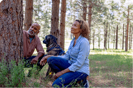 Husband and wife kneeling beside their memorial tree with their dog in a protected forest, symbolizing love, remembrance, and togetherness in nature.
